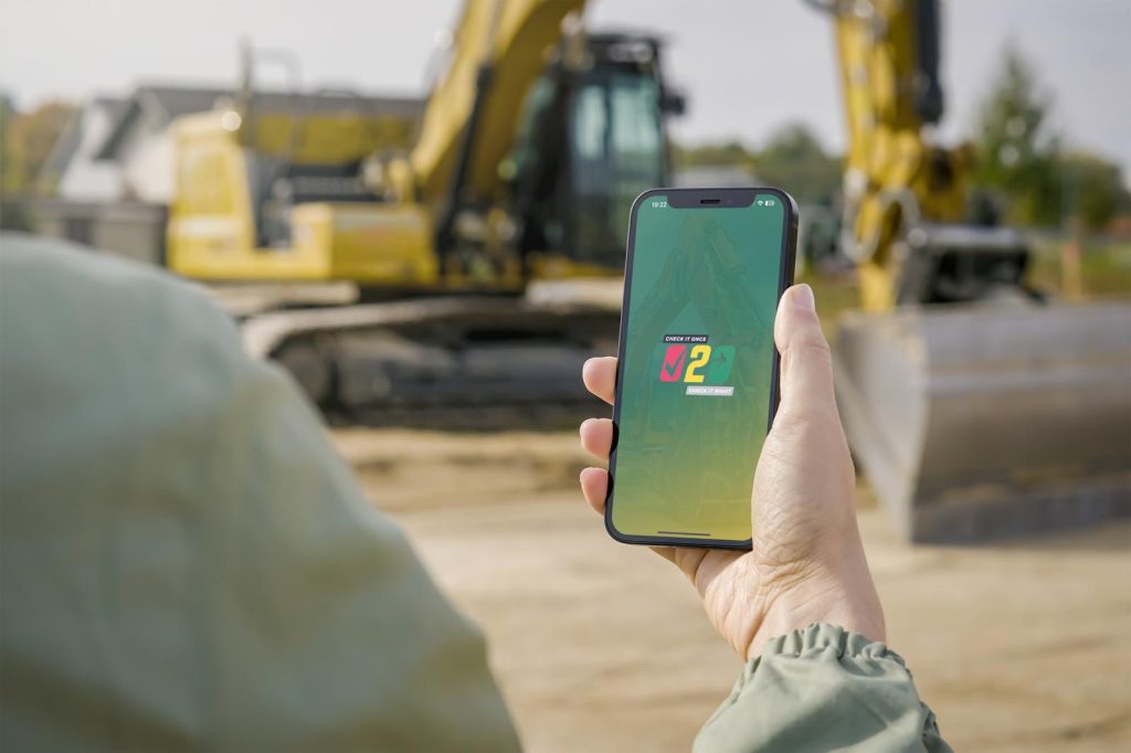 Person holding a phone with the Comply2Go app open, standing near a construction excavator on a job site.